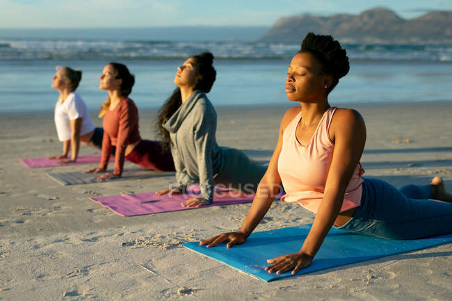 Groupe d'amis féminines diverses pratiquant le yoga, à la plage posée et amidonnante avec les yeux fermés. mode de vie sain et actif, forme physique extérieure et bien-être. — Photo de stock