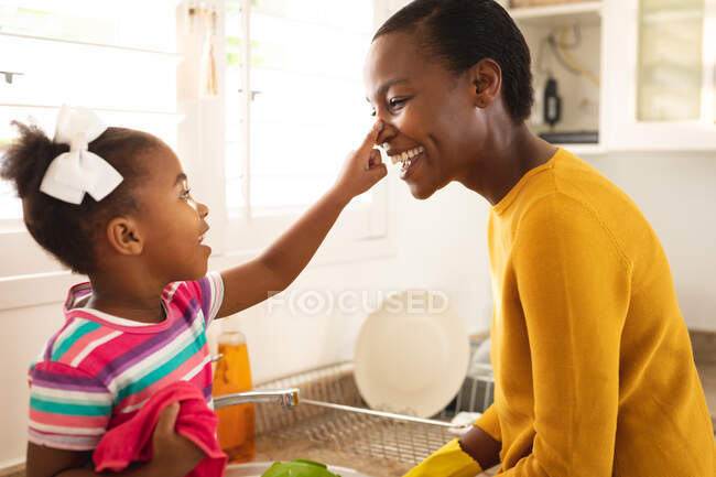 Улыбающиеся африканская американская мать и дочь веселятся на кухне, моясь вместе. Семья проводит время вместе дома. — стоковое фото