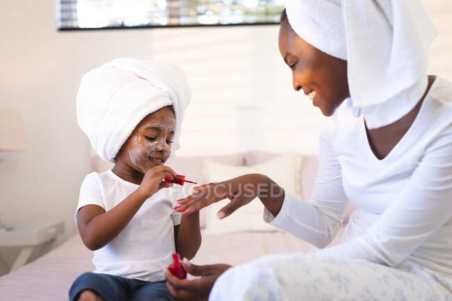 Glückliche afrikanisch-amerikanische Mutter und Tochter relaxen im Schlafzimmer, Tochter lackiert Mutter die Nägel. Familie verbringt Zeit zu Hause. — Stockfoto
