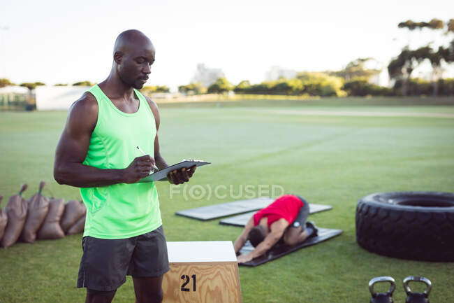 Instructor de fitness masculino afroamericano usando tableta, con colega haciendo ejercicio al aire libre. estilo de vida activo saludable, entrenamiento cruzado para fitness. - foto de stock