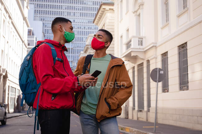 Two mixed race male friends wearing face masks and backpacks in city street using smartphone. backpacking holiday, city travel break during coronavirus covid 19 pandemic. — Stock Photo