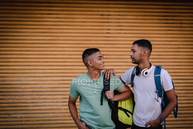Dos amigos varones de raza mixta sonrientes vistiendo mochilas mirándose y sonriendo. vacaciones de mochilero, escapada a la ciudad. - foto de stock