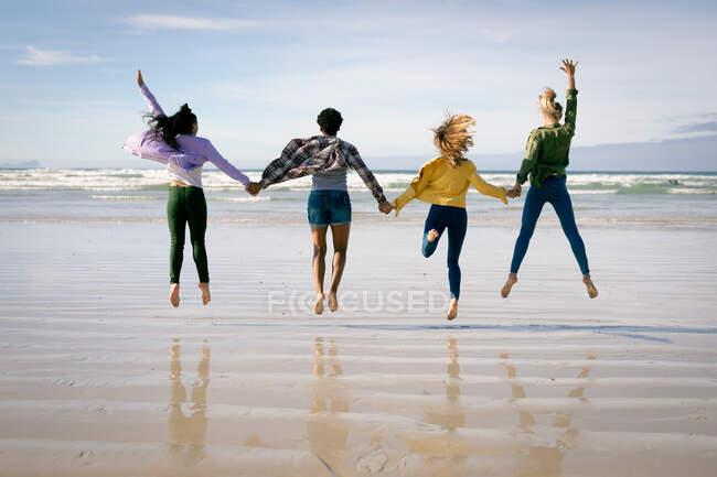 Happy group of diverse female friends having fun, walking along beach holding hands and jumping. holiday, freedom and leisure time outdoors. — Stock Photo