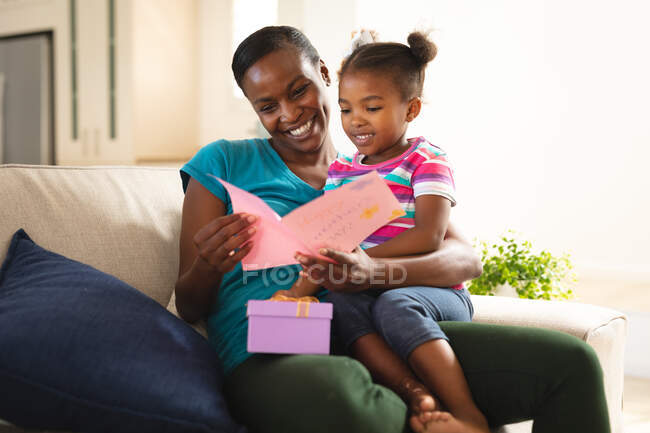 Счастливая африканская американская мать и дочь сидят на диване с карточкой и подарком от дочери. Семья проводит время вместе дома. — стоковое фото