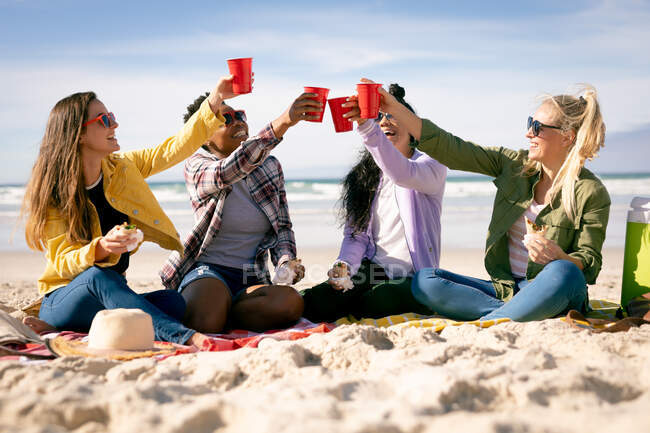 Fröhliche Gruppe unterschiedlicher Freundinnen, die Spaß haben, am Strand sitzen und anstoßen. Freundinnen-Bonding am Strand. — Stockfoto