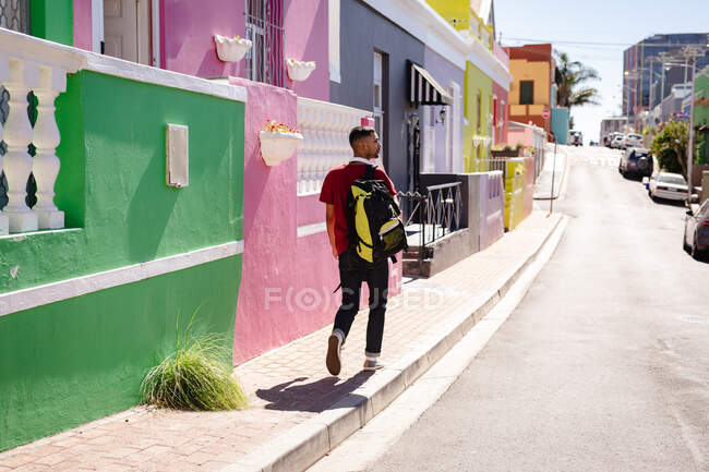 Вид ззаду мішаного гоночного чоловіка з рюкзаком, що йде різнокольоровим розписом, сонячною вулицею міста. канікули в рюкзаку, перерва на проїзд по місту . — стокове фото