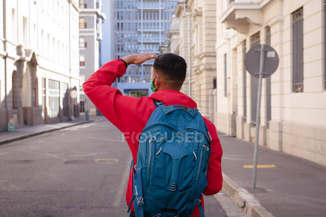 Rear view of mixed race man wearing face mask and backpack standing in city street. backpacking holiday, city travel break during coronavirus covid 19 pandemic. — Stock Photo