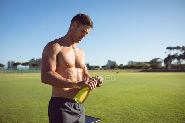 Кавказский мускулистый мужчина без рубашки, держащий воду, отдыхающий во время тренировок на открытом воздухе. здоровый активный образ жизни, кросс тренировки для фитнеса. — стоковое фото