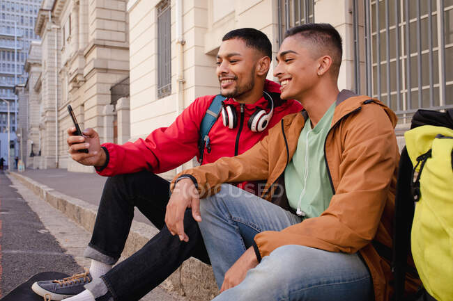 Dos amigos varones de raza mixta felices con mochilas sentadas en la calle de la ciudad tomando selfie. vacaciones de mochilero, escapada a la ciudad. - foto de stock