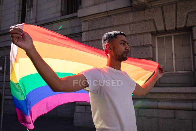 Retrato del hombre gay de raza mixta sonriente parado en la calle soleada sosteniendo la bandera del arco iris. manifestantes por la igualdad de derechos y justicia en marcha de manifestación. - foto de stock