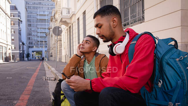 Дві змішані раси друзі-чоловіки з рюкзаками сидять на міській вулиці, один використовує смартфон. канікули в рюкзаку, перерва на проїзд по місту . — стокове фото