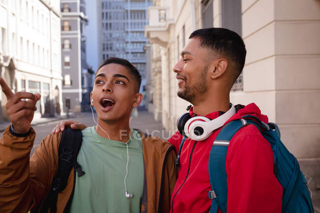 Dos amigos varones de raza mixta felices con mochilas de pie en la calle de la ciudad hablando. vacaciones de mochilero, escapada a la ciudad. - foto de stock