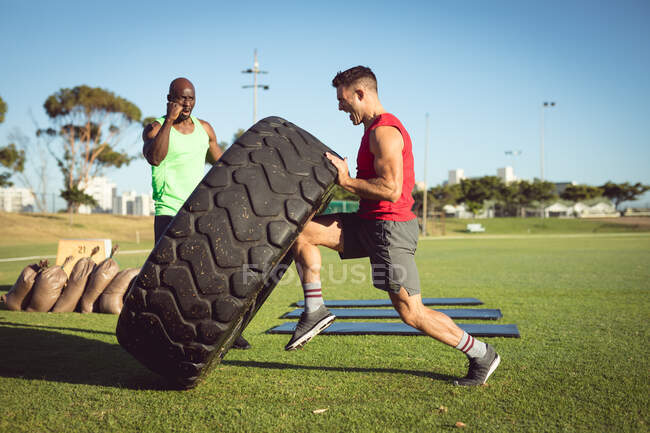 Hombre y entrenador en forma diversa haciendo ejercicio al aire libre, alentando y levantando neumáticos pesados. estilo de vida activo saludable, entrenamiento cruzado para fitness. - foto de stock