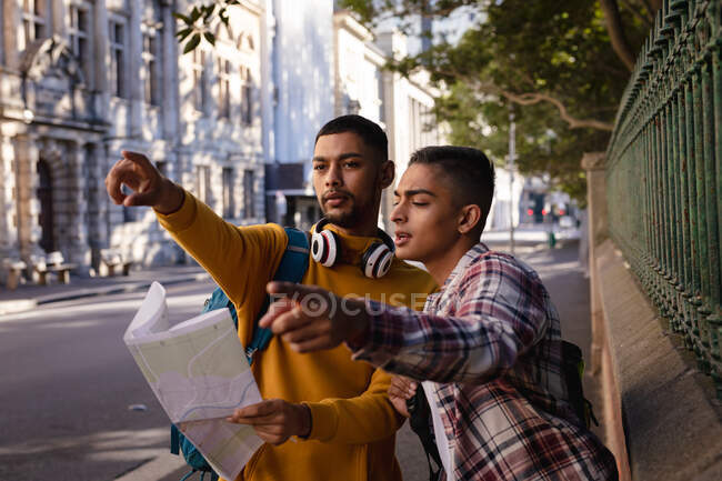 Dos amigos varones de raza mixta de pie en la calle de la ciudad, mirando el mapa, hablando y señalando la dirección. vacaciones de mochilero, escapada a la ciudad. - foto de stock