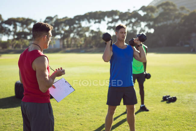 Two fit diverse men and trainer exercising outdoors, trainer timing while others lift dumbbells. healthy active lifestyle, cross training for fitness. — Stock Photo
