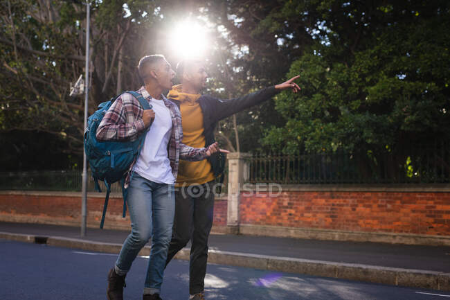 Due amici maschi di razza mista felici che portano zaini che camminano per strada, uno indicando. vacanza zaino in spalla, city travel break. — Foto stock