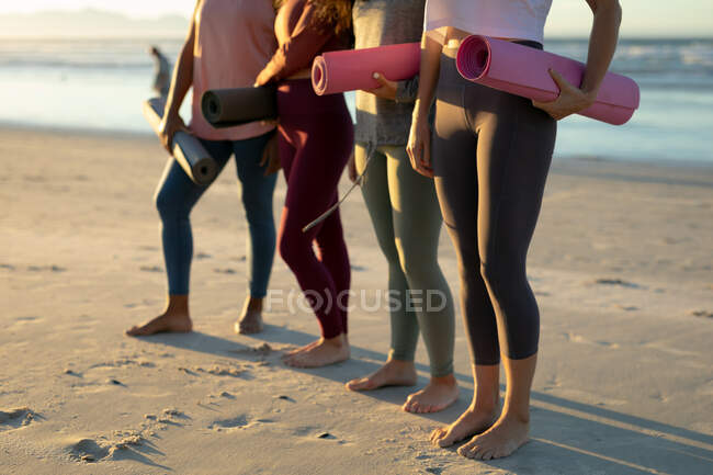 Baixa seção de diversas amigas praticando ioga, na praia segurando tapetes de ioga. estilo de vida ativo saudável, fitness ao ar livre e bem-estar. — Fotografia de Stock