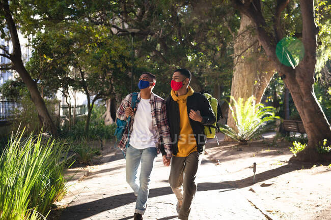 Two mixed race male friends wearing face masks and backpacks walking in city park. backpacking holiday, city travel break during coronavirus covid 19 pandemic. — Stock Photo