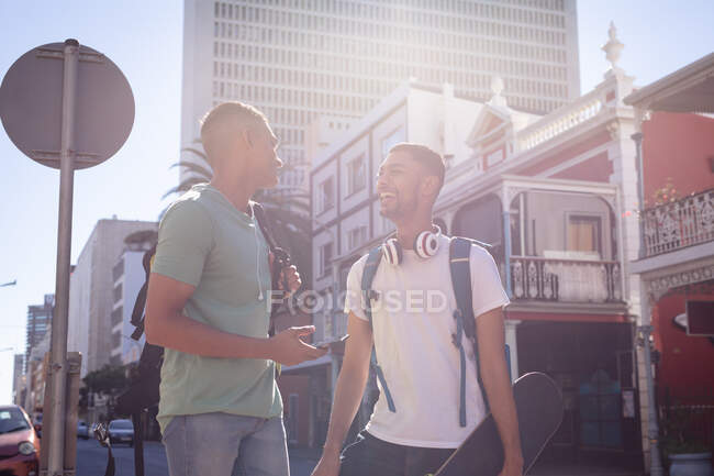 Dos felices amigos varones de raza mixta con mochilas paradas en la soleada calle de la ciudad hablando. vacaciones de mochilero, escapada a la ciudad. - foto de stock