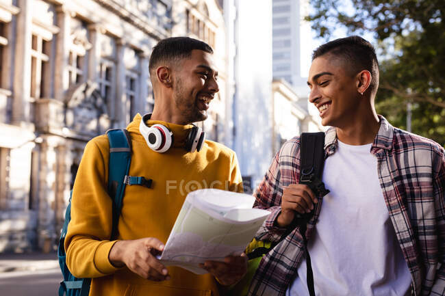 Двоє щасливих змішаних друзів чоловічої статі, що стоять на міській вулиці з рюкзаками, дивляться на карту і розмовляють. канікули в рюкзаку, перерва на проїзд по місту . — стокове фото