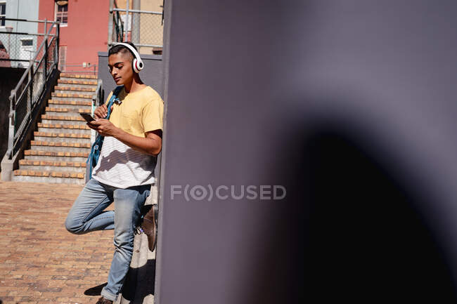 Lächelnder Mischlingsrennfahrer mit Kopfhörern, der in der sonnigen Stadtstraße mit dem Smartphone steht. Rucksackurlaub, Städtereise. — Stockfoto