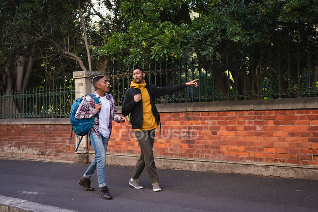 Dos amigos varones de raza mixta felices llevando mochilas caminando por la calle de la ciudad hablando, uno señalando. vacaciones de mochilero, escapada a la ciudad. - foto de stock