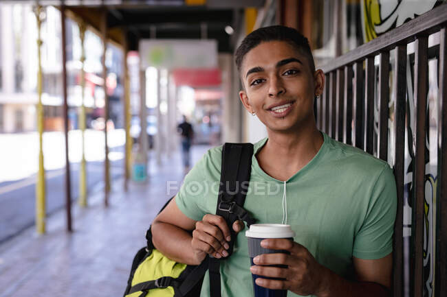 Retrato de homem de raça mista sorridente com mochila em pé na rua segurando takeaway café. mochila férias, city travel break. — Fotografia de Stock