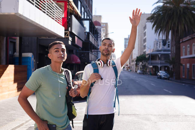 Two happy mixed race male friends with backpacks standing in sunny city street hailing taxi. backpacking holiday, city travel break. — Stock Photo