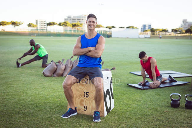 Retrato de homem caucasiano em forma de sorriso se exercitando ao ar livre, fazendo uma pausa, sentado na caixa. estilo de vida ativo saudável, treinamento cruzado para fitness. — Fotografia de Stock