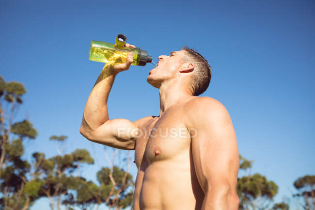 Kaukasischer muskulöser, hemdloser Mann, der Wasser trinkt, während er im Freien Sport macht. gesunder aktiver Lebensstil, Crosstraining für Fitness. — Stockfoto