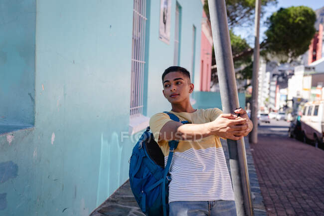 Homem de raça mista feliz usando mochila em pé na ensolarada rua da cidade balançando no poste de luz. mochila férias, city travel break. — Fotografia de Stock