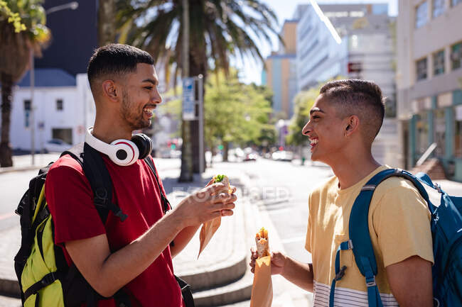 Two smiling mixed race male friends with backpacks standing in sunny city street eating fast food. backpacking holiday, city travel break. — Stock Photo