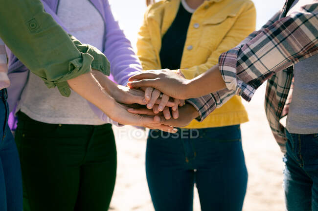 Sección media de mujeres que apilan las manos contra el cielo azul. amigas unidas en la playa. - foto de stock