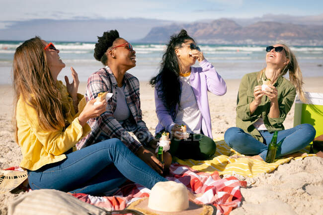 Feliz grupo de diversas amigas divirtiéndose, sentadas en la playa y comiendo. amigas unidas en la playa. - foto de stock