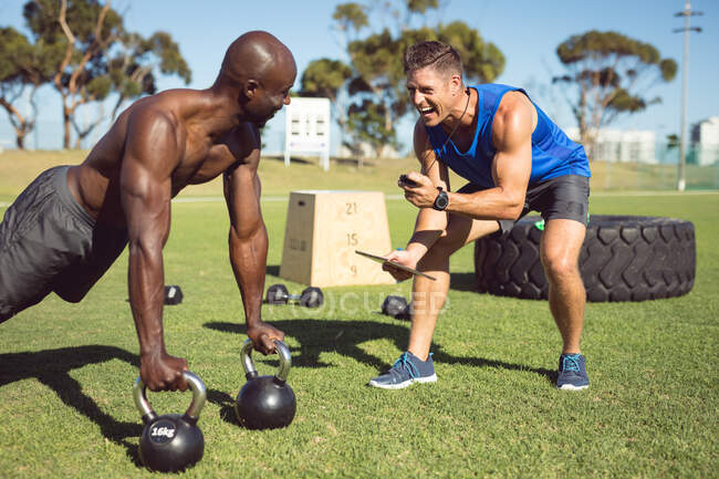 African american muscular man exercising outdoors with kettle bells and fitness instructor. healthy active lifestyle, cross training for fitness. — Stock Photo