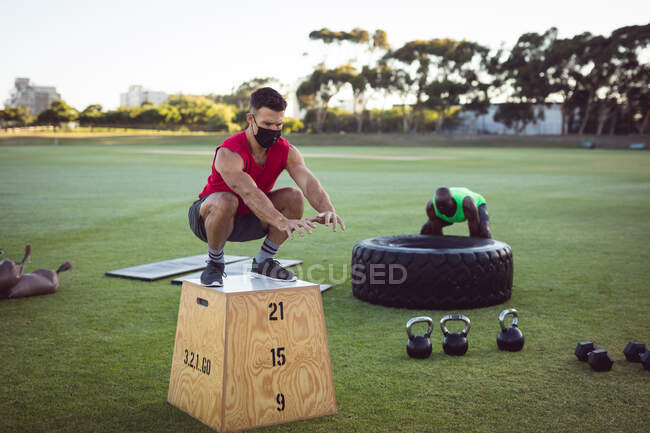 Fit caucasian man wearing face mask exercising outdoors, jumping onto box. healthy active lifestyle, cross training for fitness during coronavirus covid 19 pandemic. — Stock Photo