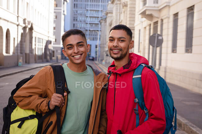 Retrato de dos amigos varones de raza mixta sonrientes con mochilas en la calle de la ciudad. vacaciones de mochilero, escapada a la ciudad. - foto de stock
