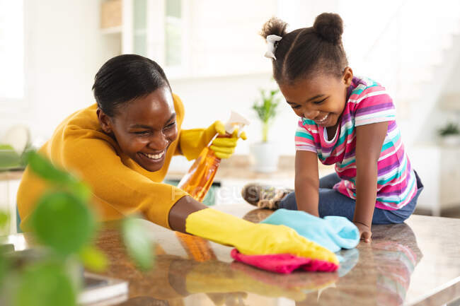 Улыбающиеся африканские американские мать и дочь веселятся, убирая кухонный стол вместе. Семья проводит время вместе дома. — стоковое фото