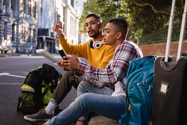Дві змішані раси друзі чоловічої статі сидять на вулиці з багажем, використовуючи смартфон і дивлячись на карту. канікули в рюкзаку, перерва на проїзд по місту . — стокове фото