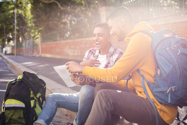 Zwei fröhliche Mischlingsfreunde sitzen mit Rucksäcken auf der Straße, lachen, schauen auf die Karte. Rucksackurlaub, Städtereise. — Stockfoto