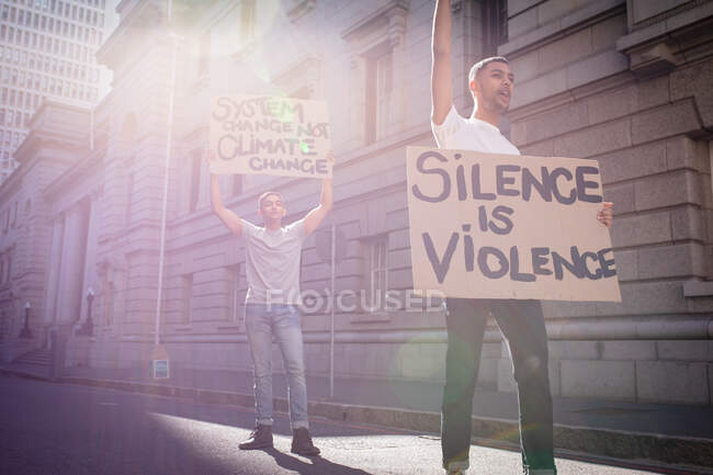 Dos amigos varones de raza mixta llevando pancartas de protesta con consignas en la soleada calle de la ciudad. manifestantes por la igualdad de derechos y justicia manifestándose en la ciudad. - foto de stock