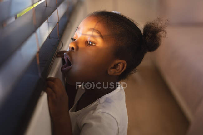 Surpreendida menina afro-americana de pé e olhando através de persianas em um dia ensolarado. passar tempo livre em casa. — Fotografia de Stock