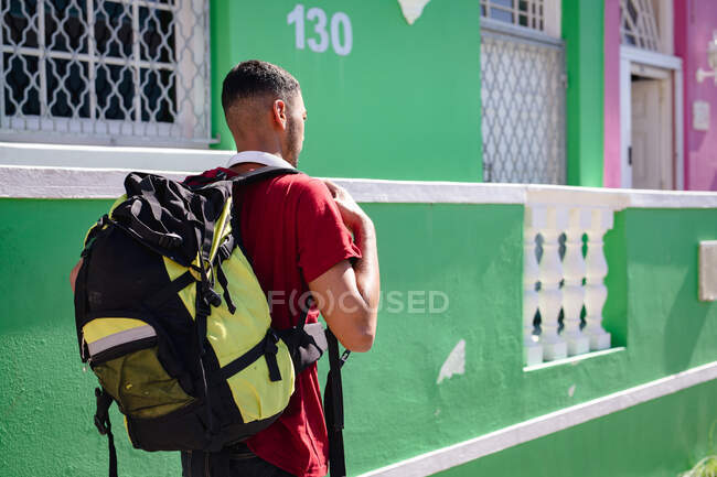 Rear view of mixed race man carrying backpack walking in sunny city street. backpacking holiday, city travel break. — Stock Photo