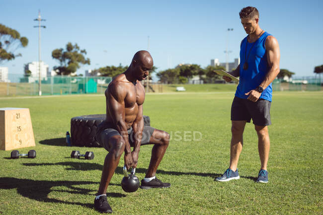 Uomo muscoloso afroamericano che si allena all'aperto con campane e istruttore di fitness. sano stile di vita attivo, cross training per il fitness. — Foto stock
