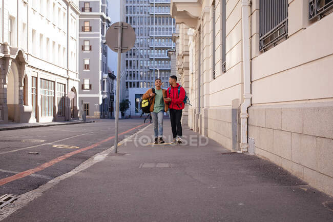 Zwei lächelnde männliche Freunde gemischter Rassen, die Rucksäcke tragen und in der Stadtstraße miteinander reden. Rucksackurlaub, Städtereise. — Stockfoto