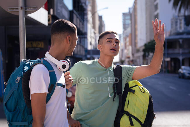 Dos amigos varones de raza mixta con mochilas de pie en la soleada calle de la ciudad, uno llamando al taxi. vacaciones de mochilero, escapada a la ciudad. - foto de stock