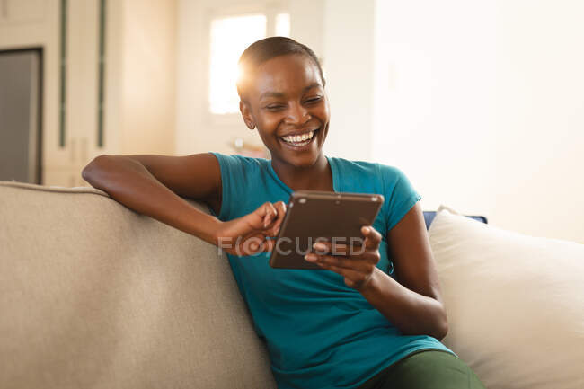 Riendo mujer afroamericana relajante, sentado en el sofá usando la tableta. pasar tiempo libre en casa. - foto de stock