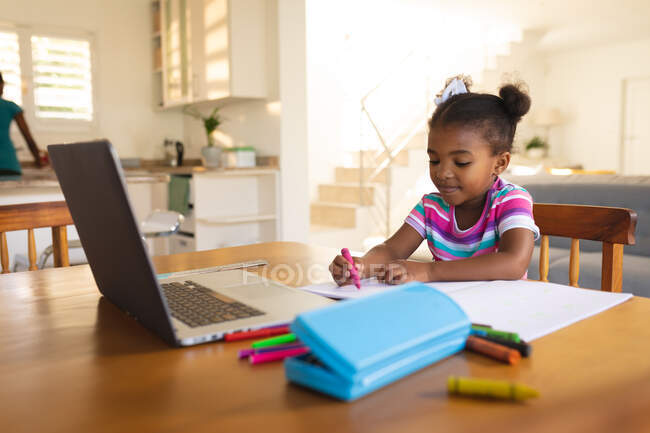 Sorrindo menina afro-americana sentada à mesa de jantar, usando laptop desenho no livro. escola online, educação em casa. — Fotografia de Stock
