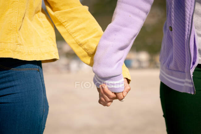 Mittelteil eines diversifizierten bisexuellen Paares, das Händchen haltend am Strand entlang geht. Freundinnen-Bonding am Strand — Stockfoto