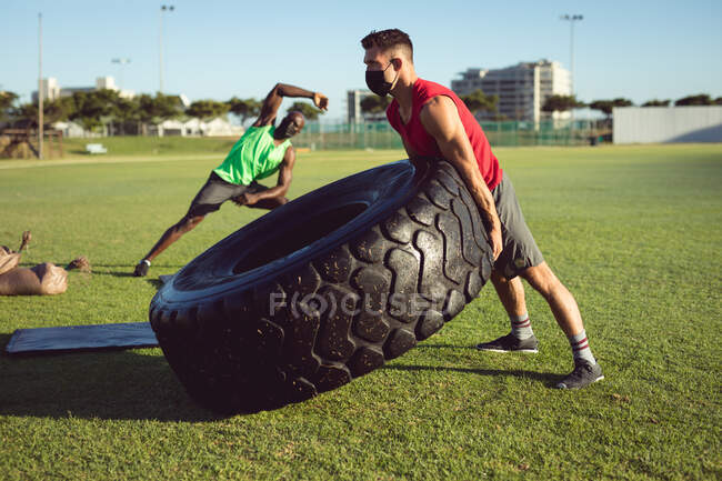 Dos hombres diversos caben ejercitando al aire libre, levantando el neumático pesado y estirando. estilo de vida activo saludable, entrenamiento cruzado para fitness. - foto de stock